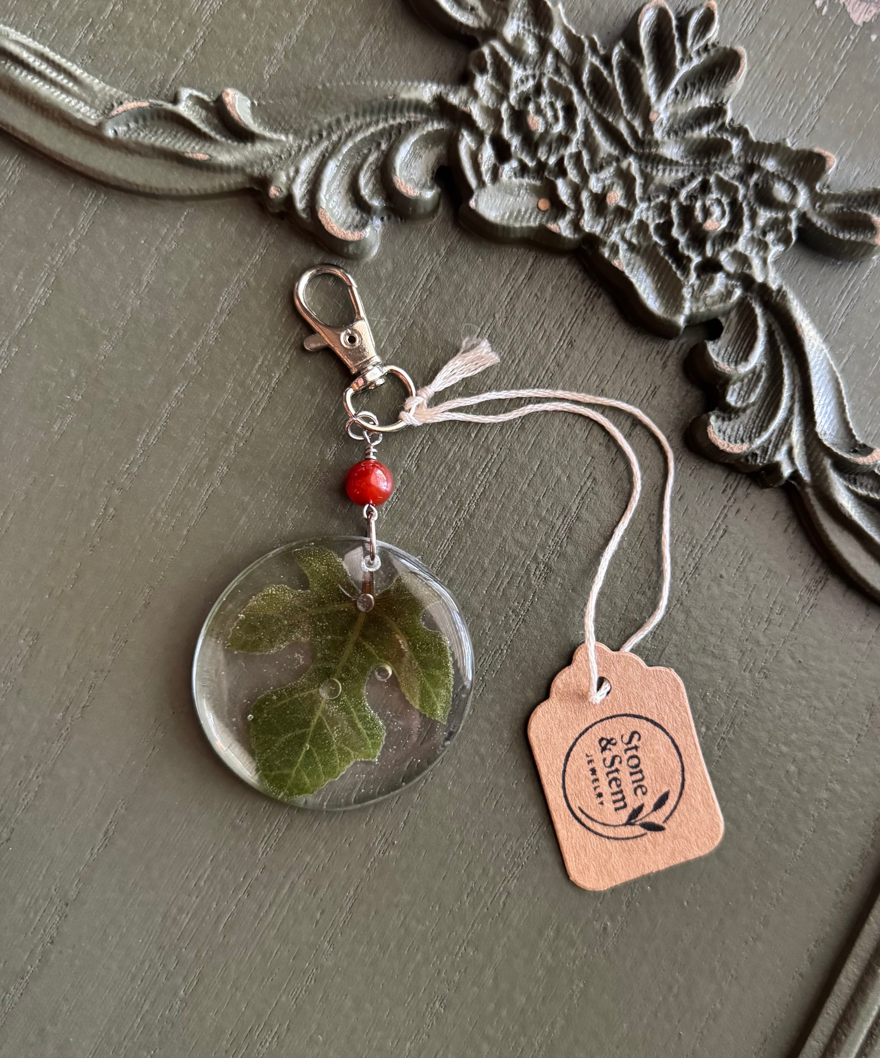 Fig leaves preserved in clear round resin keychain with red stone bead and silver-tone lobster clasp, displayed on Stone & Stem Jewelry tag