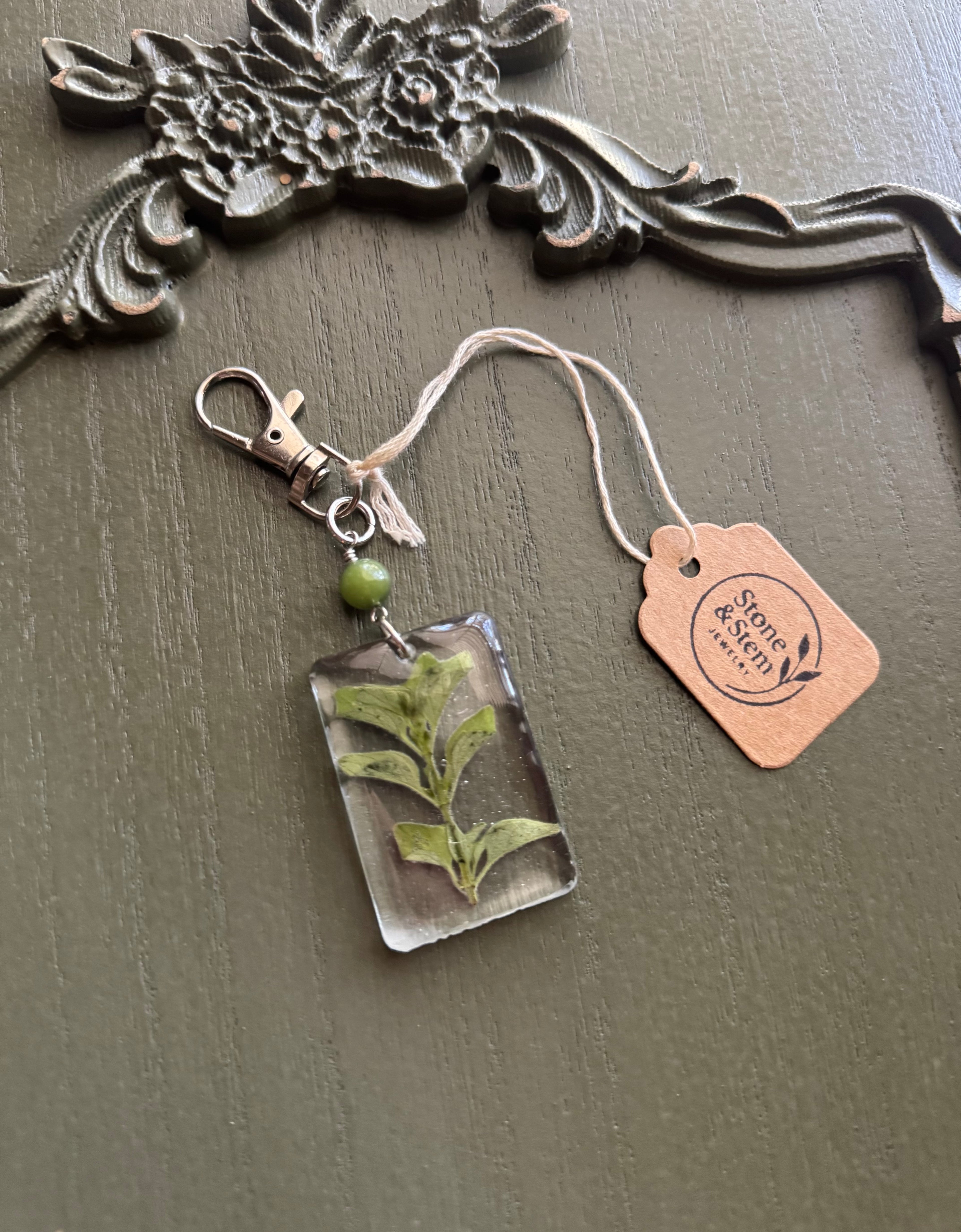 Oregano sprig preserved in clear rectangular resin keychain with green stone bead and silver-tone lobster clasp, displayed on Stone & Stem Jewelry tag