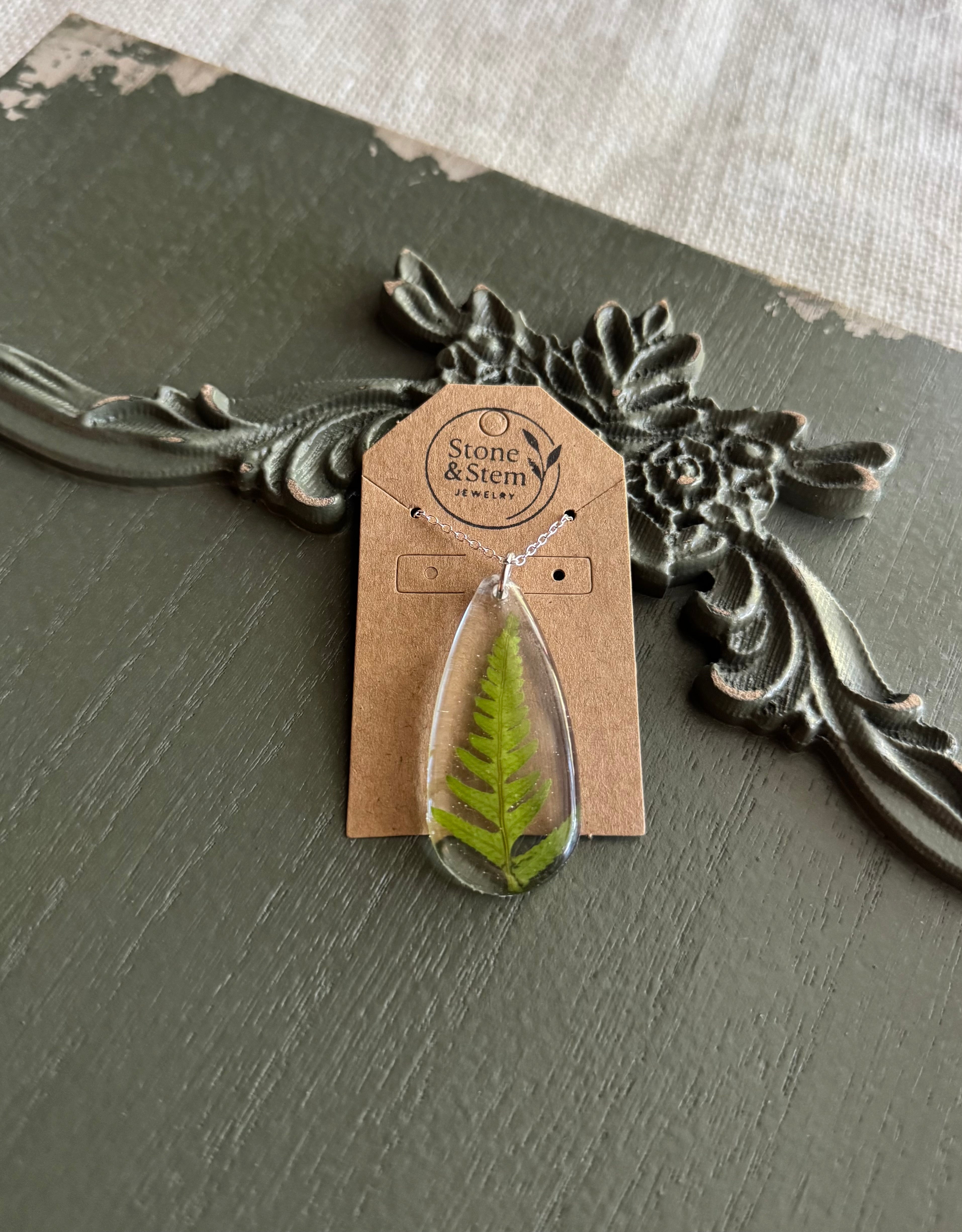Real fern frond preserved in a clear teardrop resin pendant on a silver chain, displayed on a Stone & Stem Jewelry kraft card