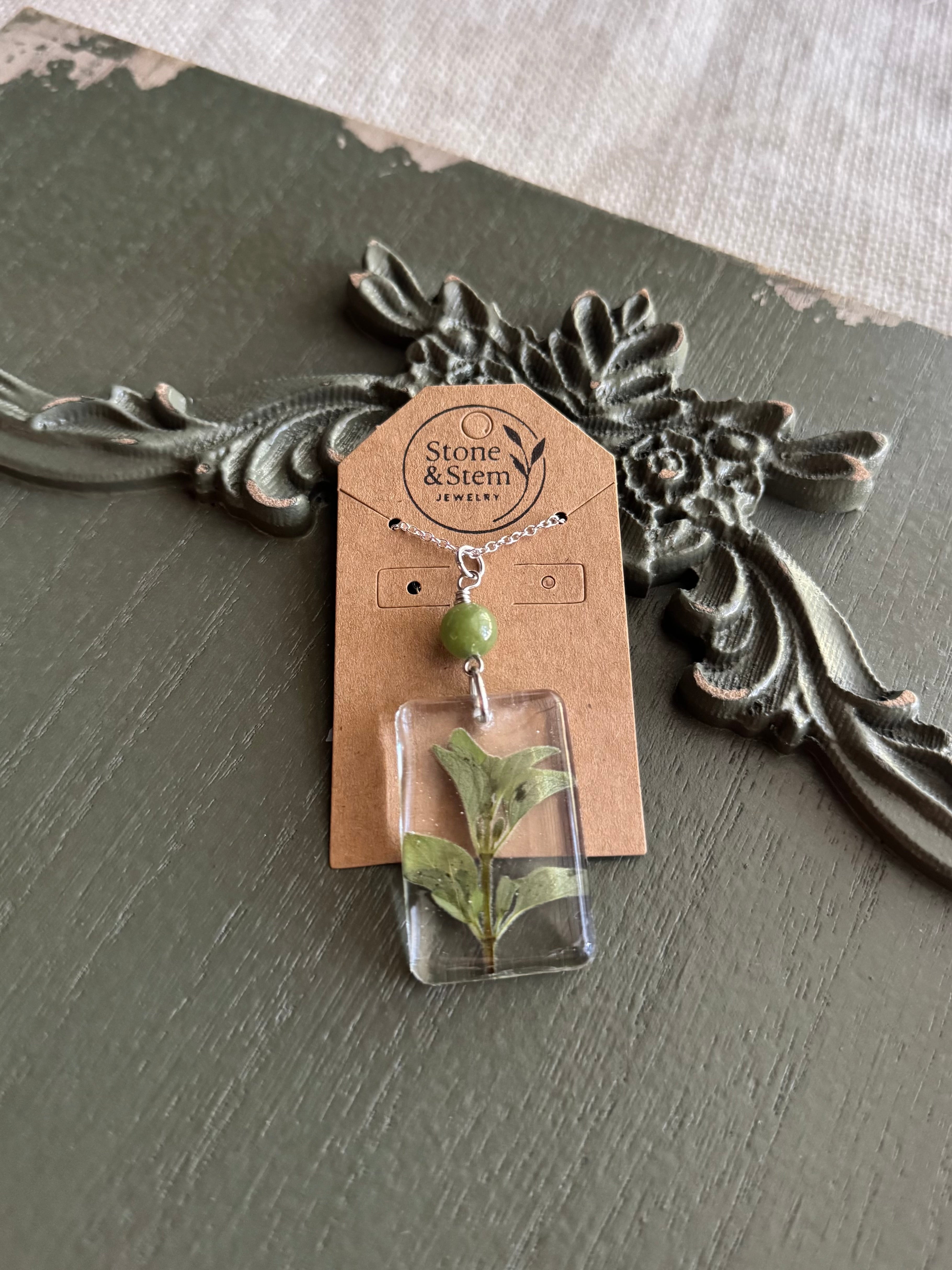 Real pressed oregano sprig in a clear square resin pendant with sage green stone bead on a silver chain, on a Stone & Stem Jewelry kraft card