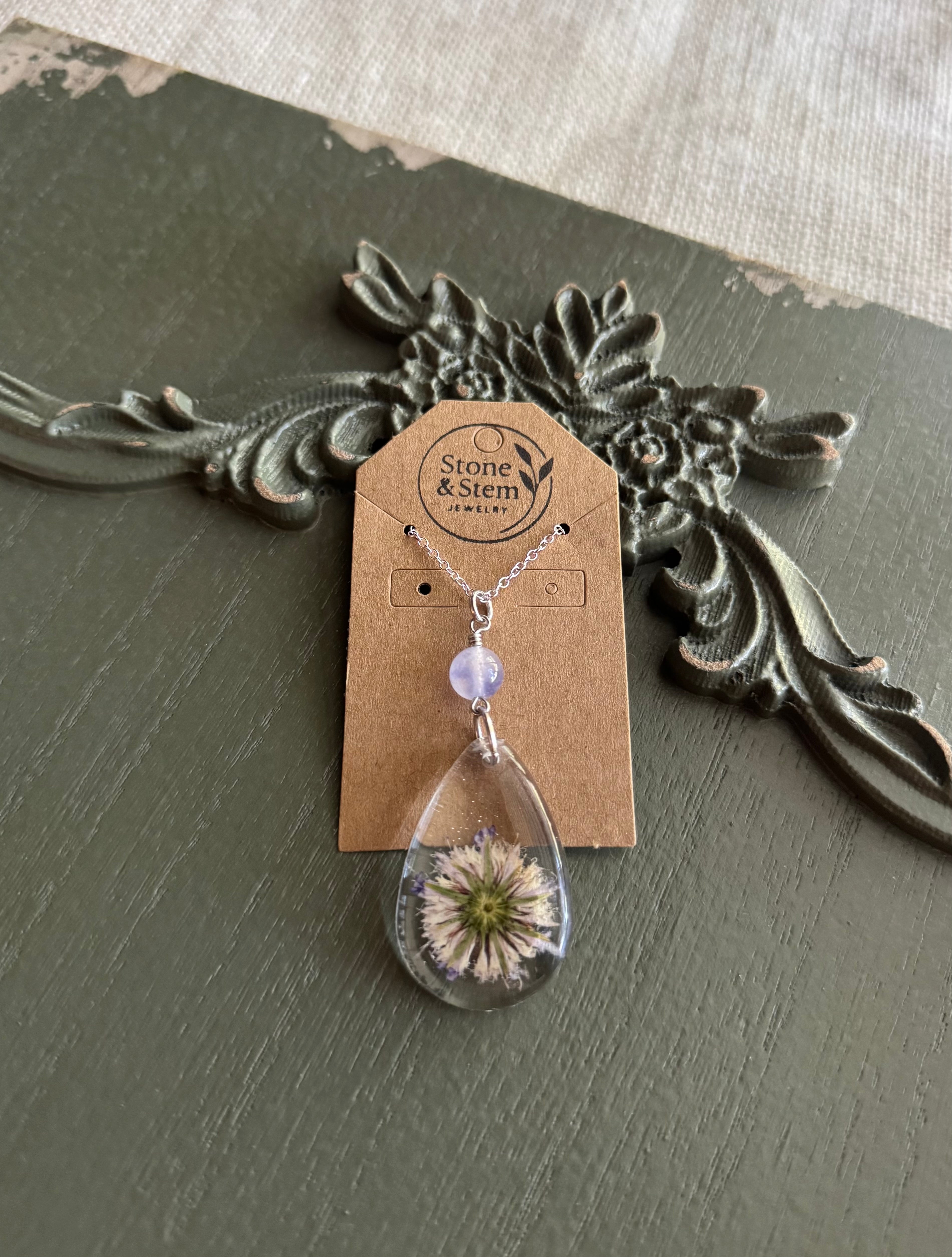 Pressed wildflower in a clear teardrop resin pendant with lavender bead on a silver chain, displayed on a Stone & Stem Jewelry kraft card
