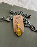 Real pressed dandelion in a clear oval resin pendant with yellow stone bead on a silver chain, displayed on a Stone & Stem Jewelry kraft card