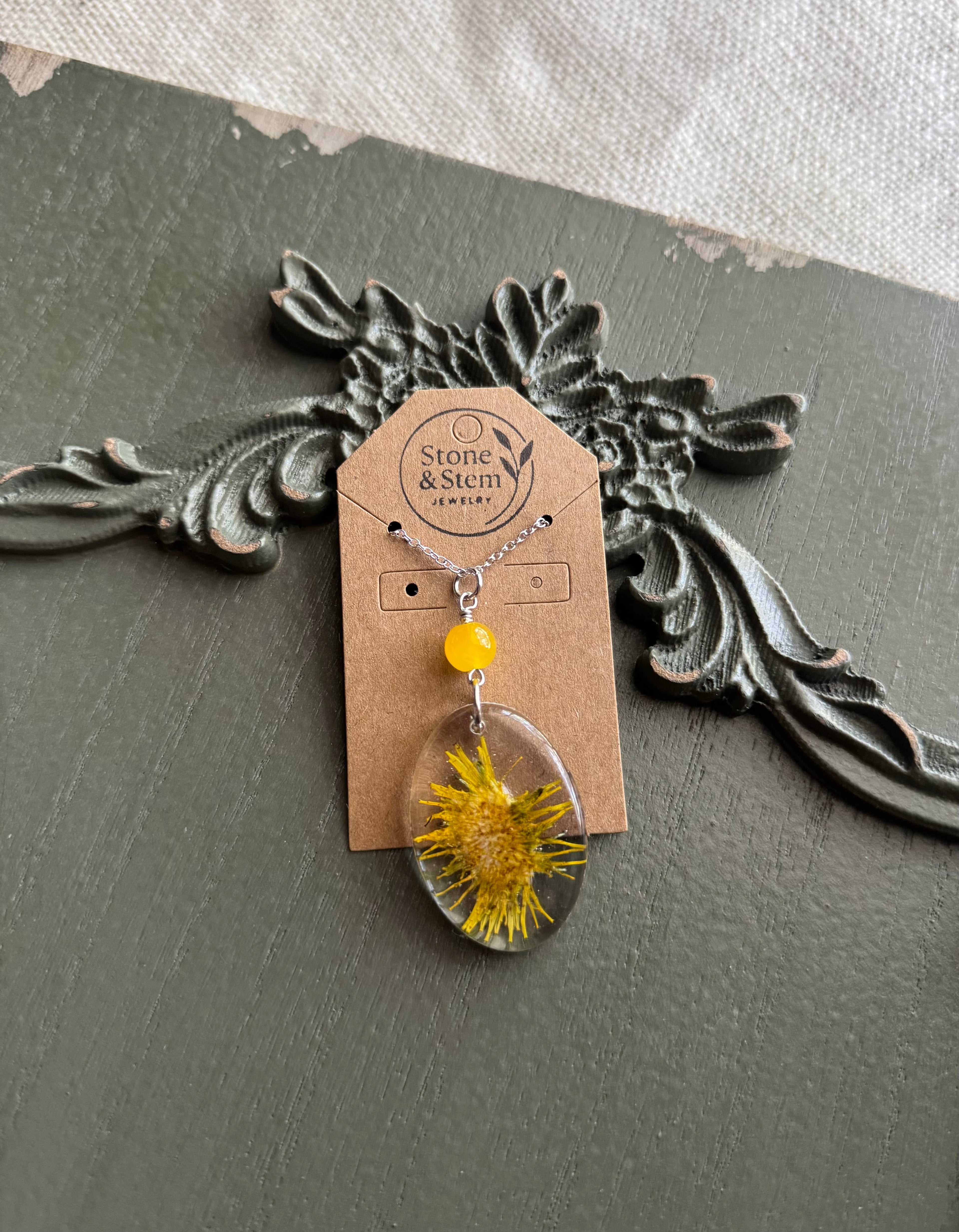 Real pressed dandelion in a clear oval resin pendant with yellow stone bead on a silver chain, displayed on a Stone & Stem Jewelry kraft card