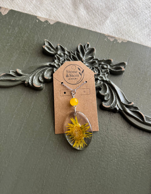 Real pressed dandelion in a clear oval resin pendant with yellow stone bead on a silver chain, displayed on a Stone & Stem Jewelry kraft card