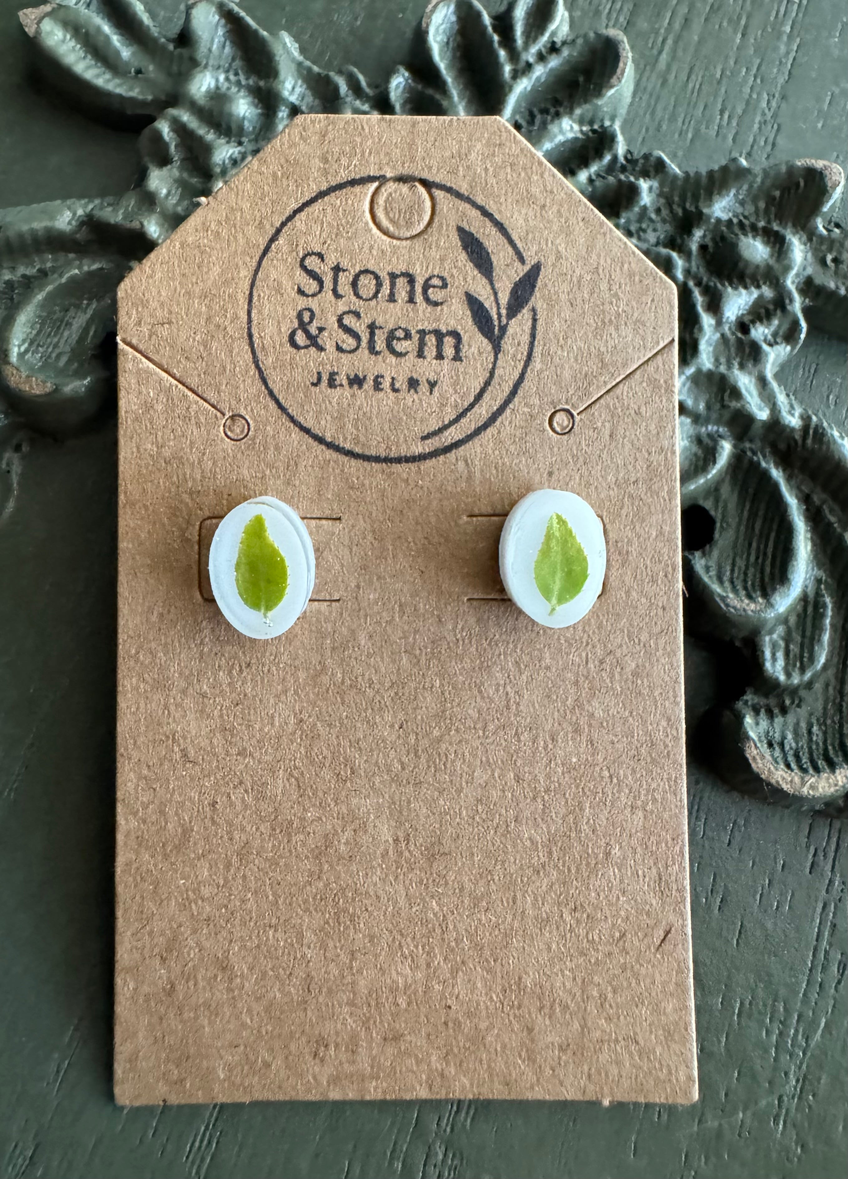 Close-up of oval white resin stud earrings showing pressed green leaf detail on a Stone & Stem Jewelry kraft card