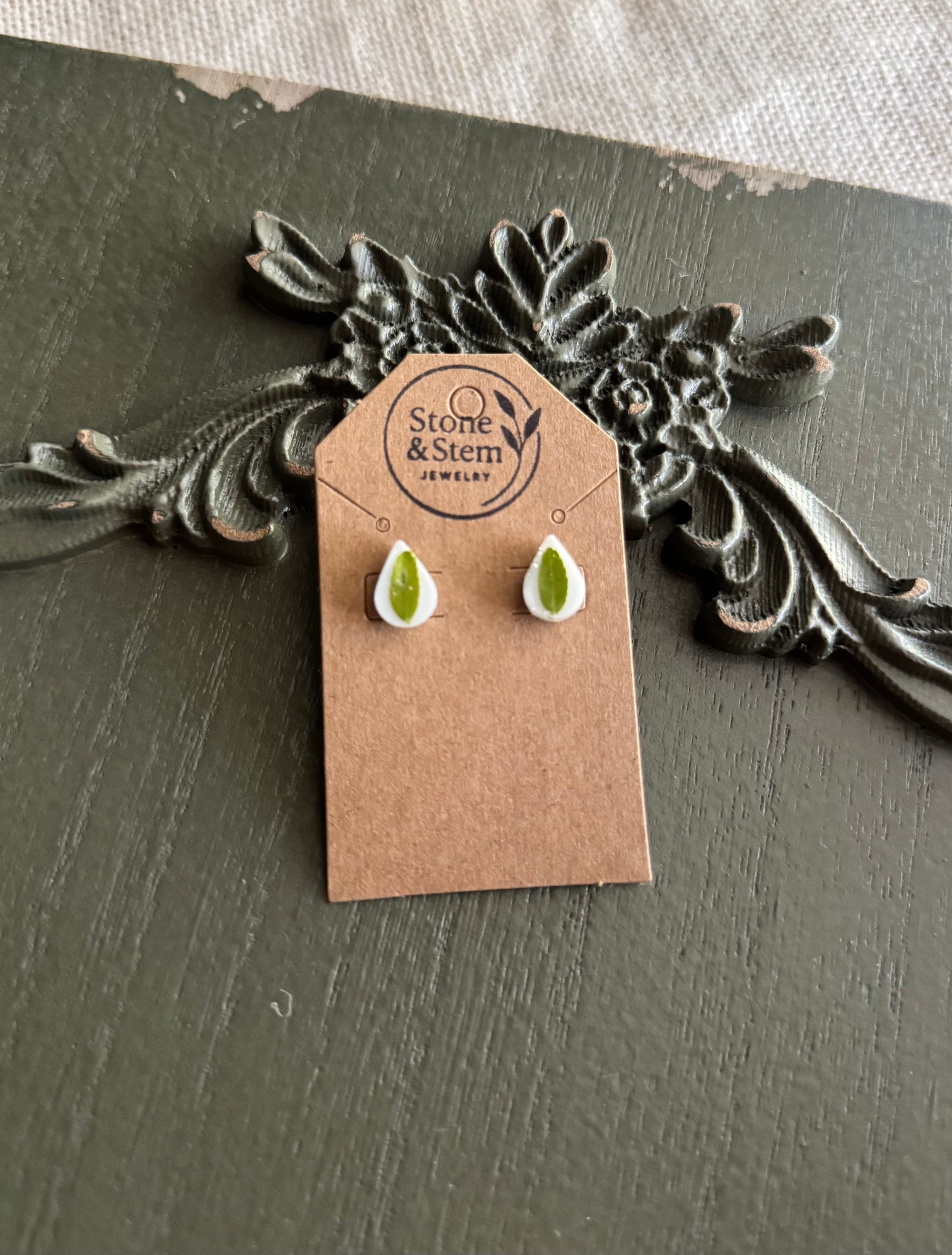 Pair of small white teardrop resin stud earrings with pressed green leaves on a Stone & Stem Jewelry kraft card