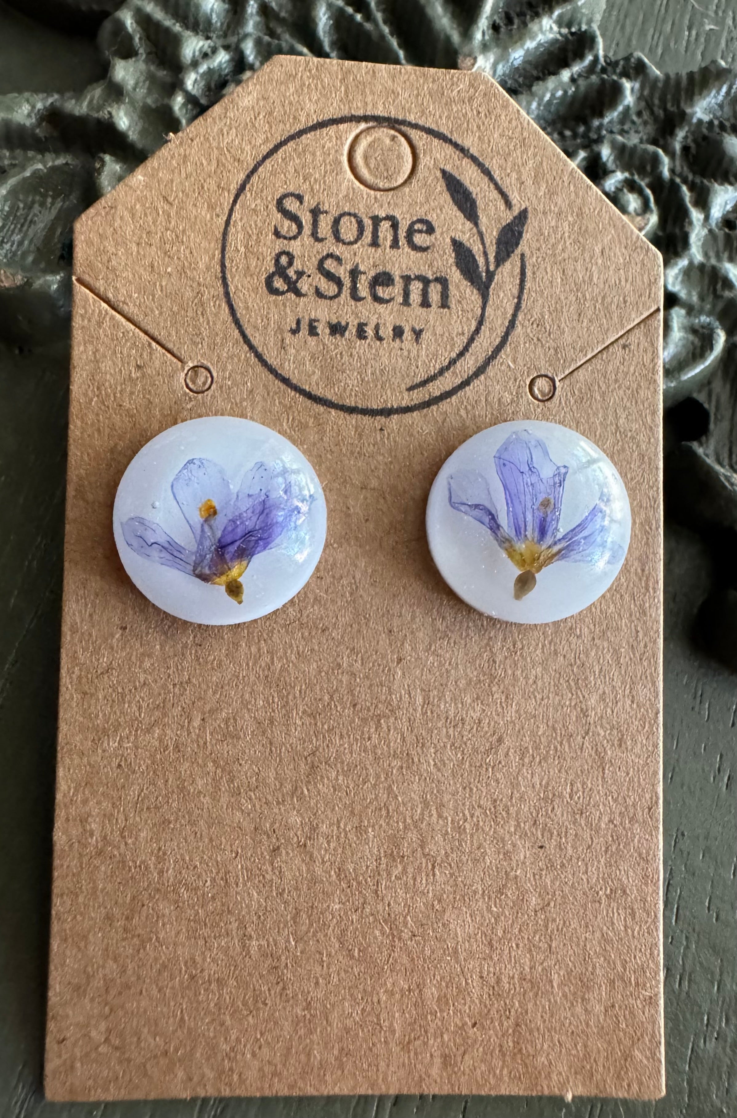 Close-up of clear round resin stud earrings showing pressed purple flower with golden center detail on a Stone & Stem Jewelry kraft card