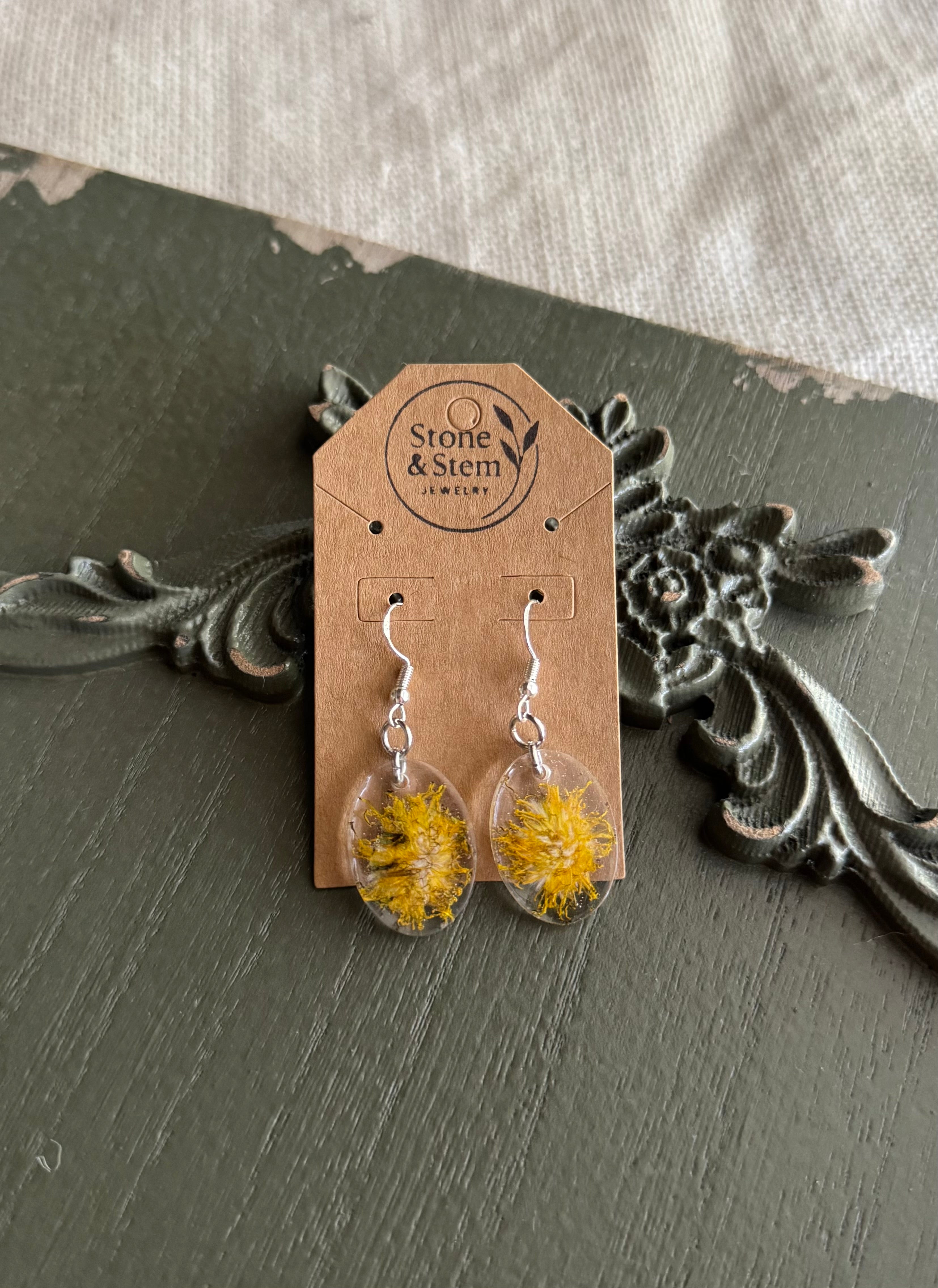 Pair of clear oval resin drop earrings with real dandelion seed heads on 925 sterling silver ear wires on a Stone & Stem Jewelry kraft card