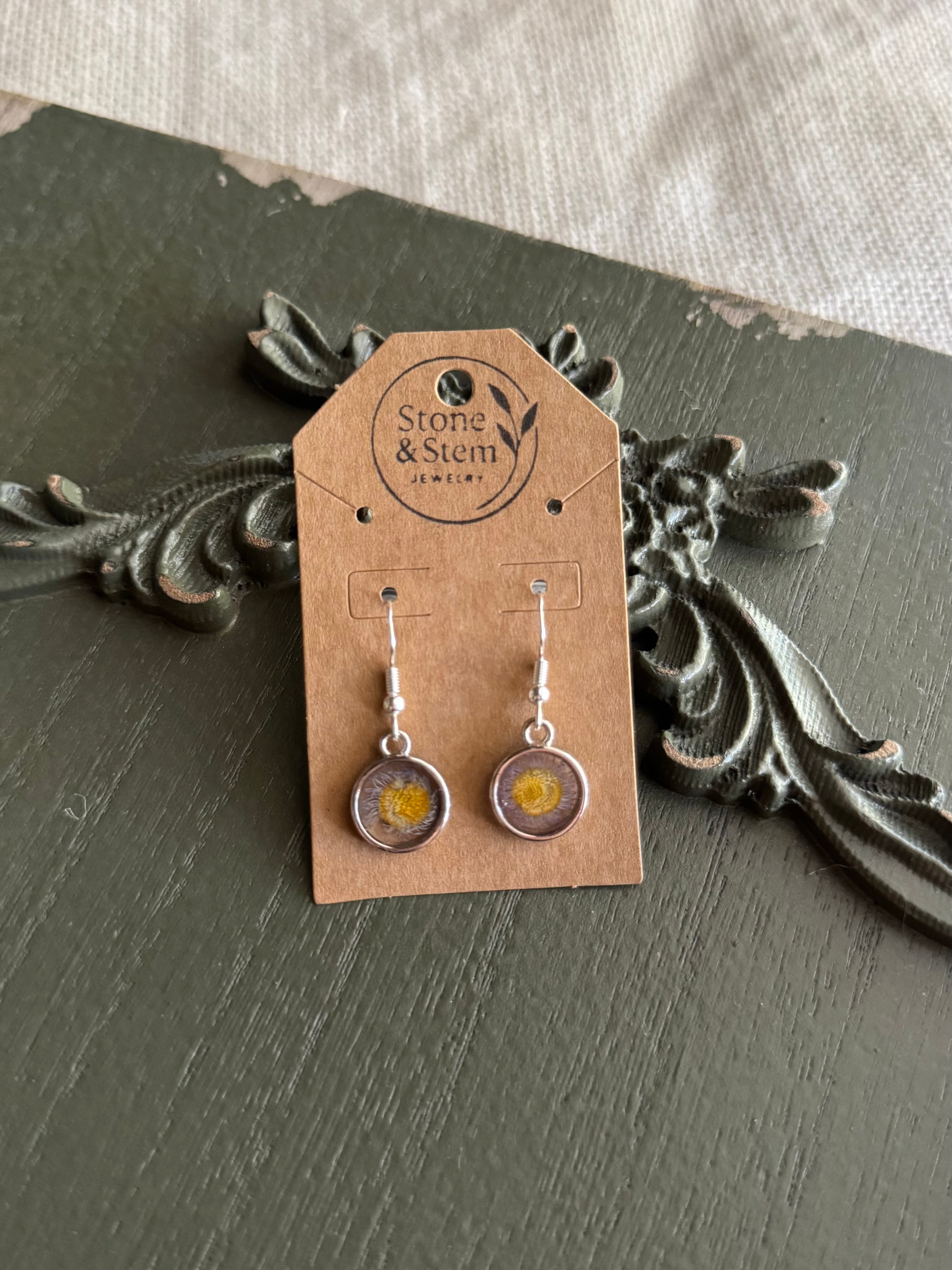 Pair of silver-framed clear round resin drop earrings with pressed purple and yellow wildflowers on 925 sterling silver ear wires on a Stone & Stem Jewelry kraft card