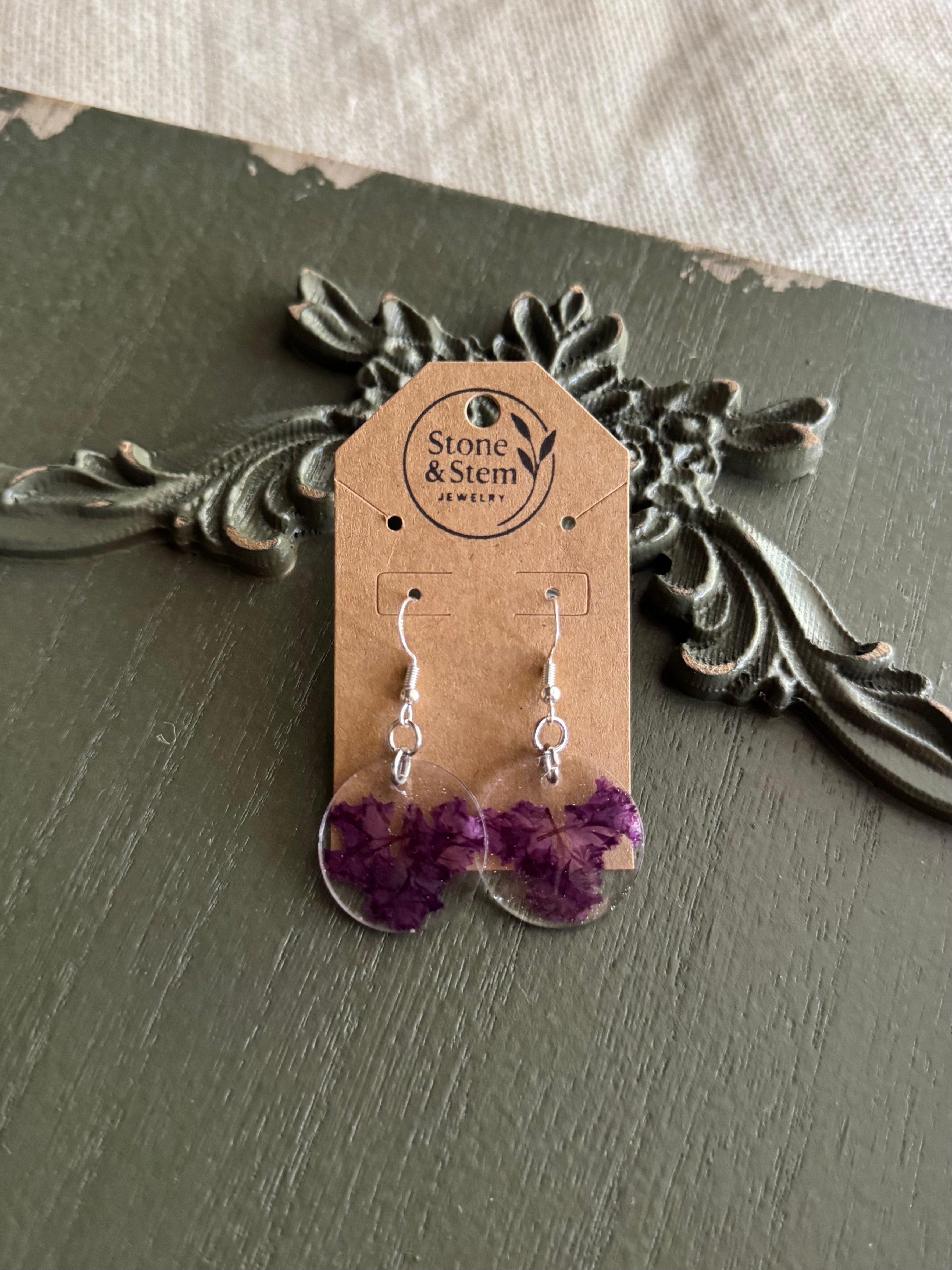 Pair of clear round resin drop earrings filled with deep purple myrtle flower petals on 925 sterling silver ear wires on a Stone & Stem Jewelry kraft card