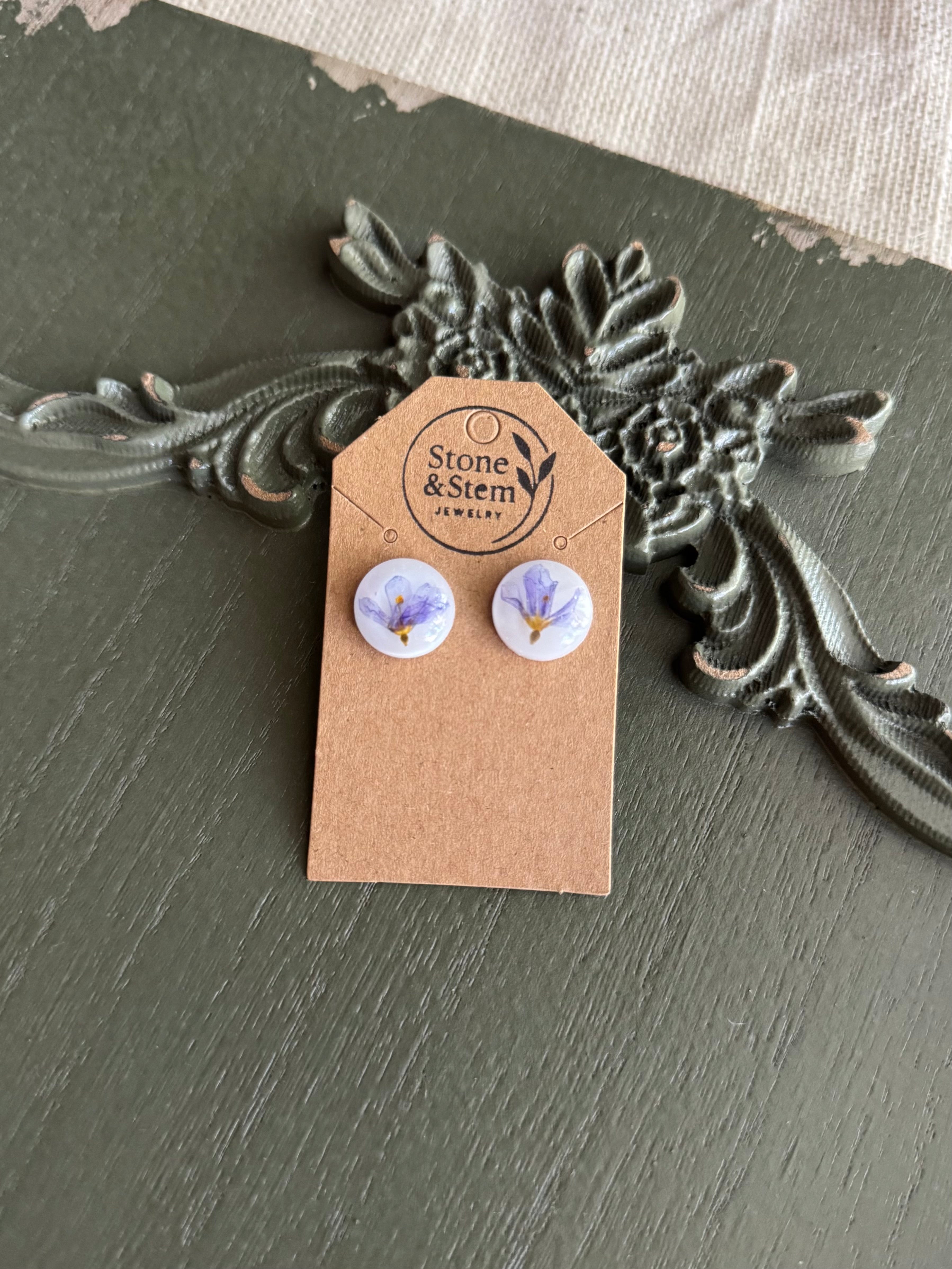 Pair of small clear round resin stud earrings with pressed purple flowers on a Stone & Stem Jewelry kraft card